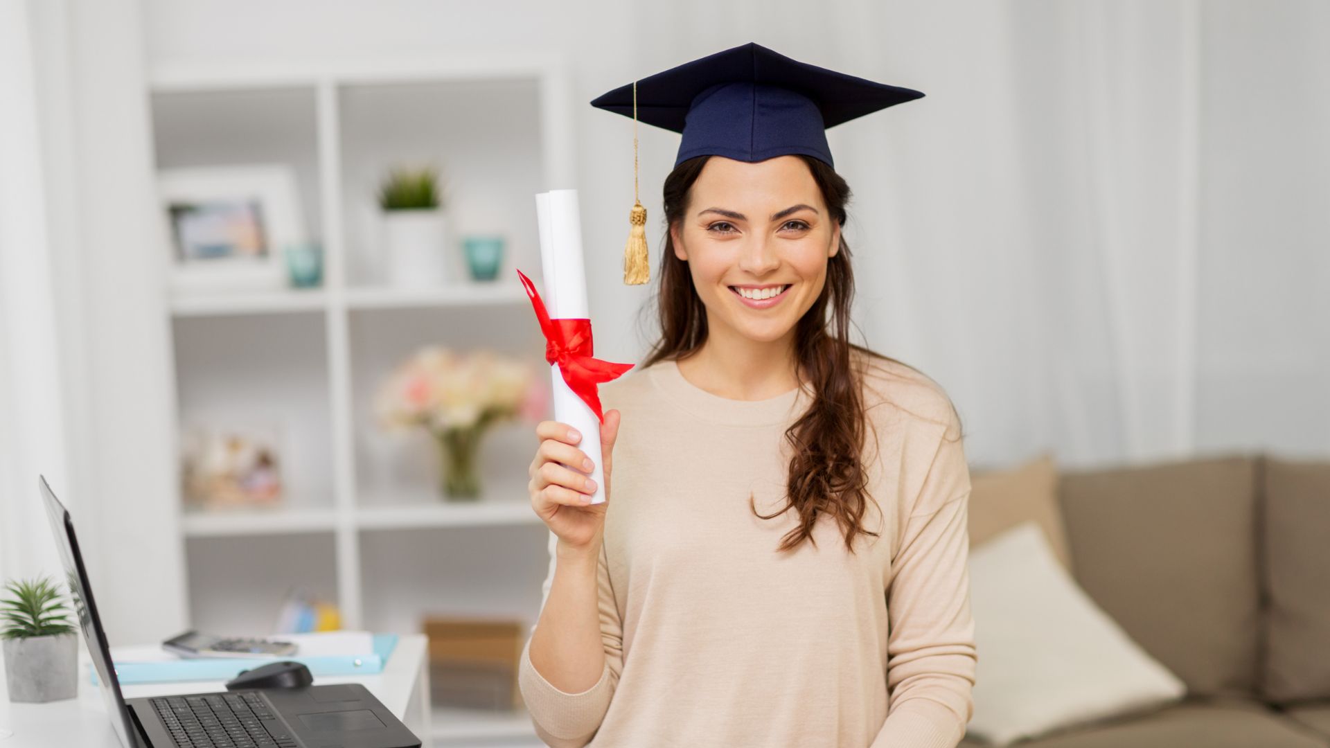 in her home office, stands a beautiful female graduate holding diploma rolled up and held together with ribbon